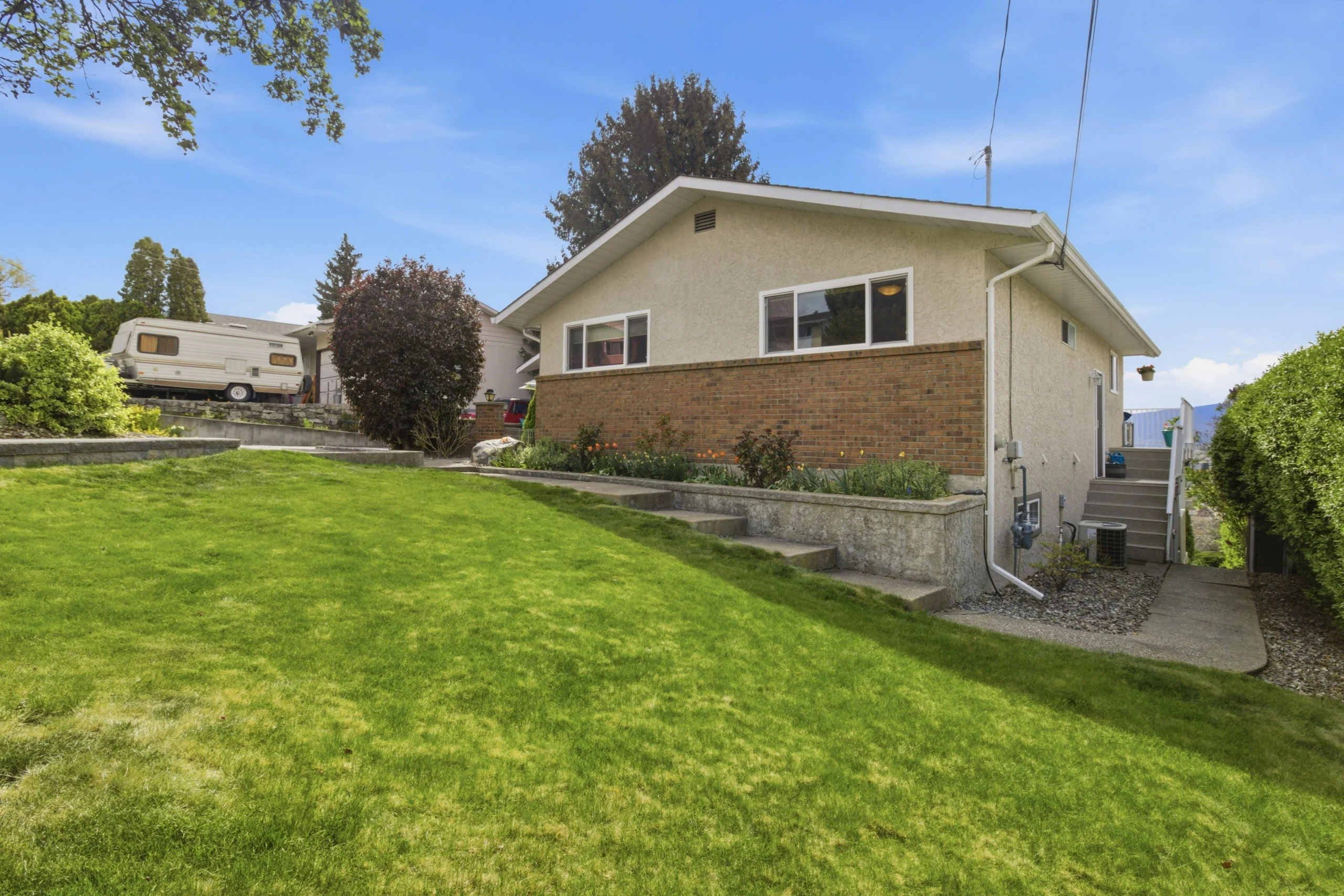 Exterior of the 869 McKenzie Rd property featuring a tan stucco and brick bungalow with a sloping green lawn and blue sky.